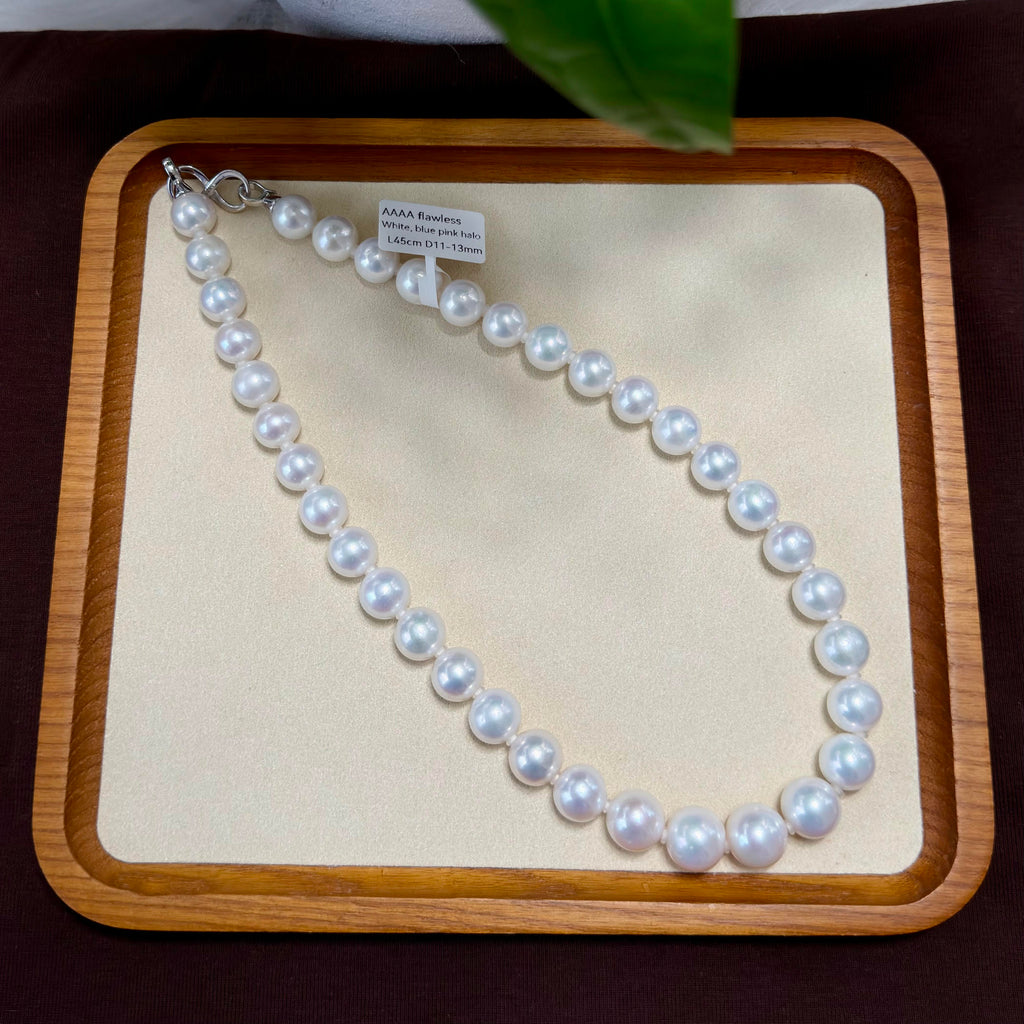 A strand of white freshwater pearls with a silver clasp, displayed on a beige insert inside a wooden tray. A small paper tag is attached to the clasp.