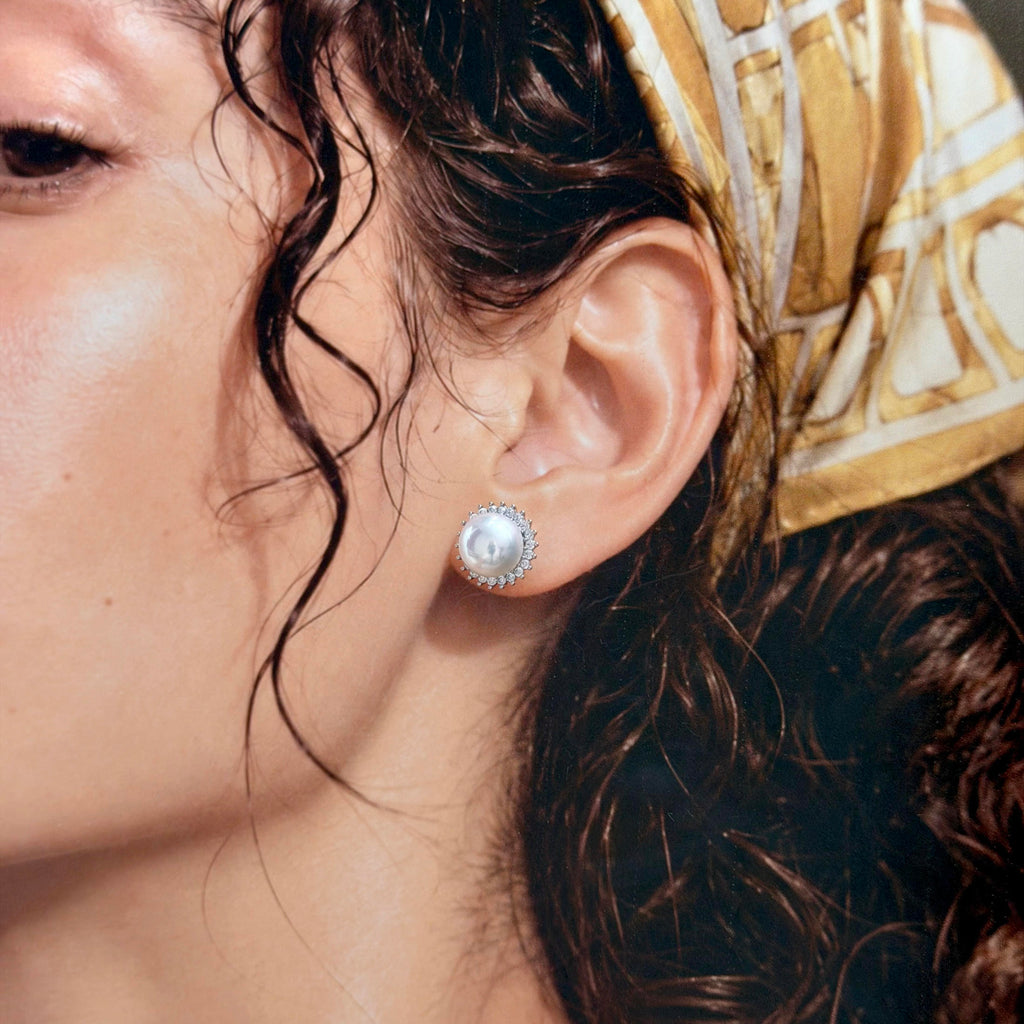 A close-up of a person's ear wearing a single round pearl stud earring with a silver-toned halo setting and a patterned gold and white headscarf.