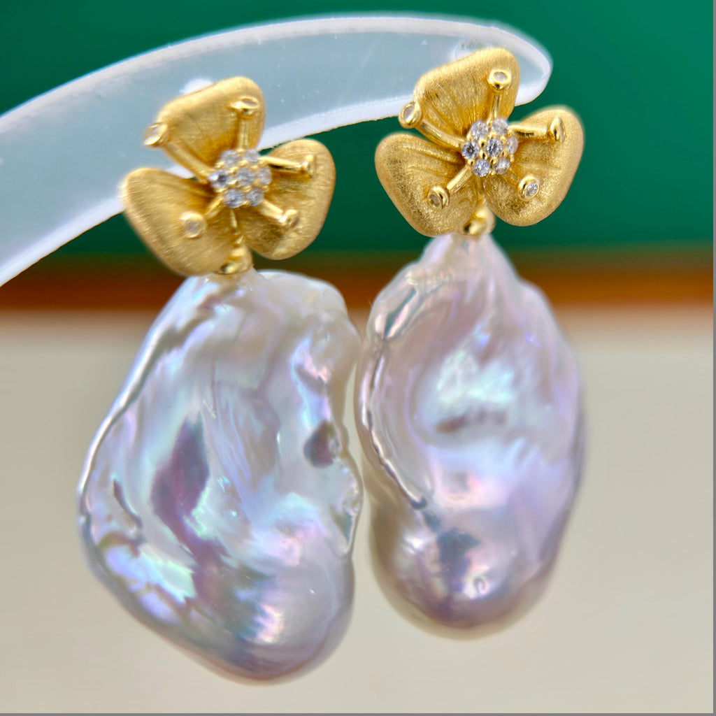 A pair of gold flower-shaped stud earrings with small clear stones in the center and large, irregular iridescent pearl drops hanging below, shown close up against a green and beige background.