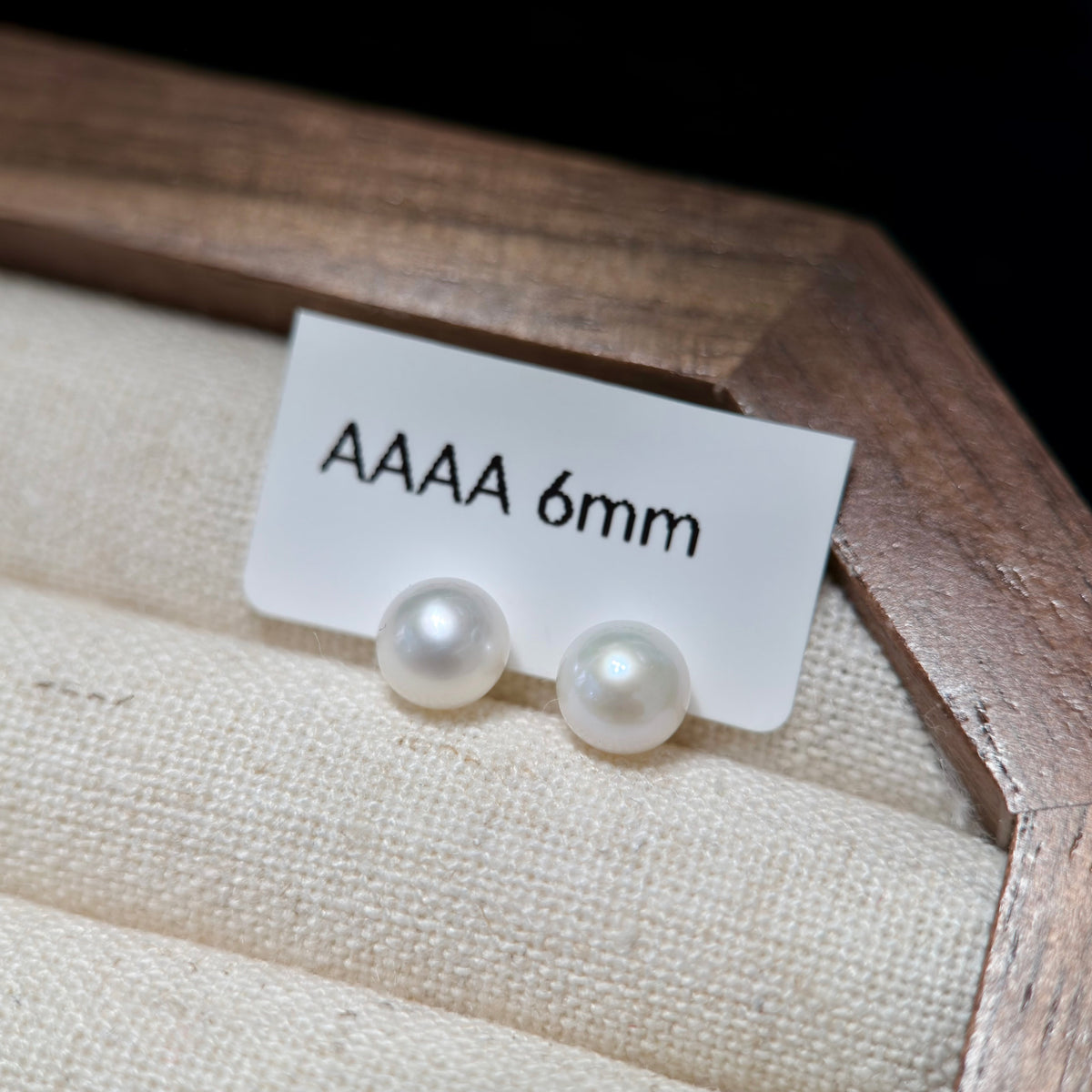 Two round white pearl stud earrings displayed on a small white card labeled 'AAAA 6mm' inside a beige fabric-lined jewelry box.