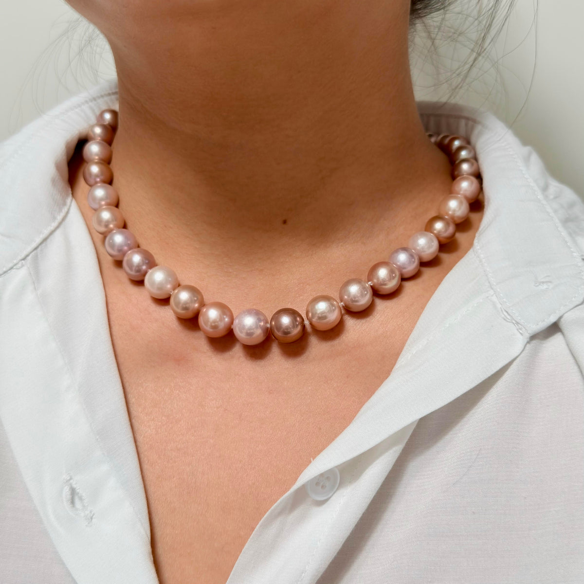 A close-up of a person wearing a strand of round pinkish pearl necklace over a white collared shirt.