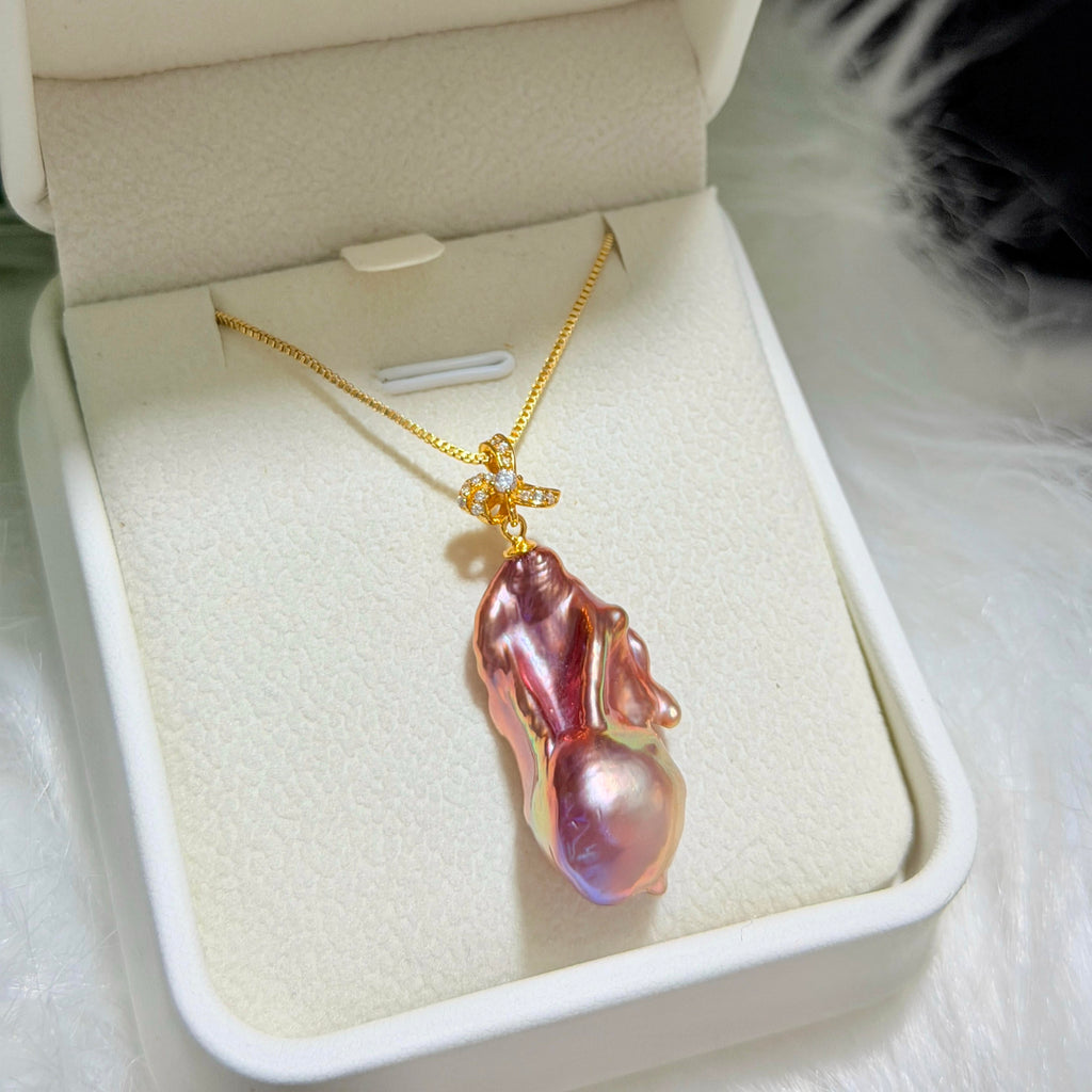 A gold necklace with a large, irregular baroque pearl pendant in a pinkish-purple iridescent finish, displayed inside an open cream-colored jewelry box.