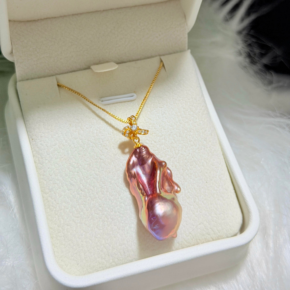 A gold necklace with a large, irregular baroque pearl pendant in a pinkish-purple iridescent finish, displayed inside an open cream-colored jewelry box.
