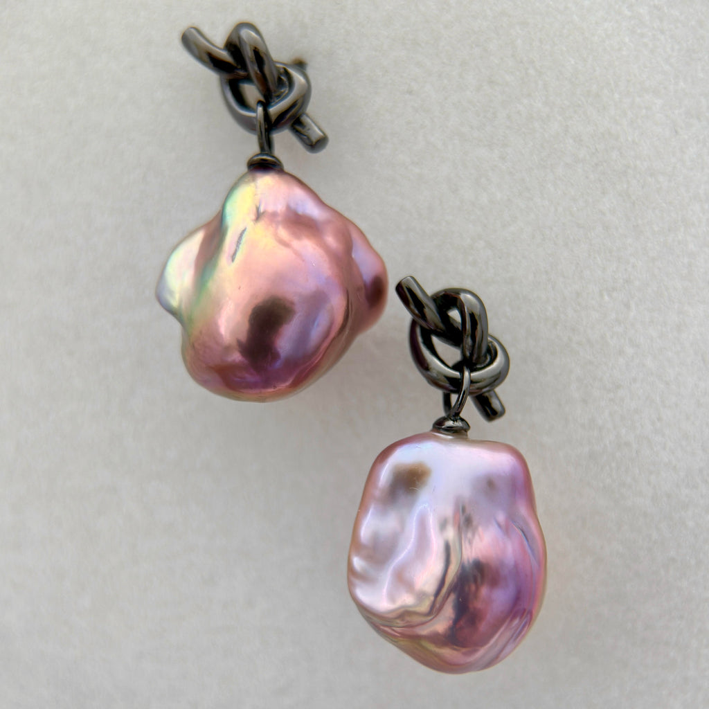 A pair of baroque pearl drop earrings with iridescent pink, purple, and green hues, suspended from dark oxidized metal chains on a light gray textured background.