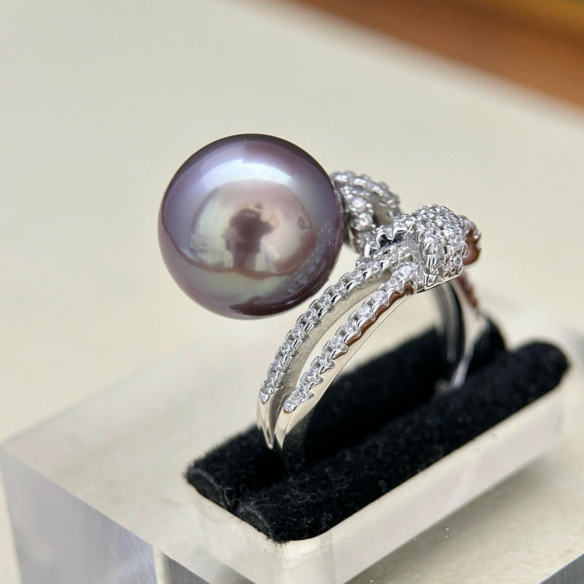A silver ring featuring a large, round, dark purple-black pearl and a snake head motif, displayed on a black velvet ring stand.