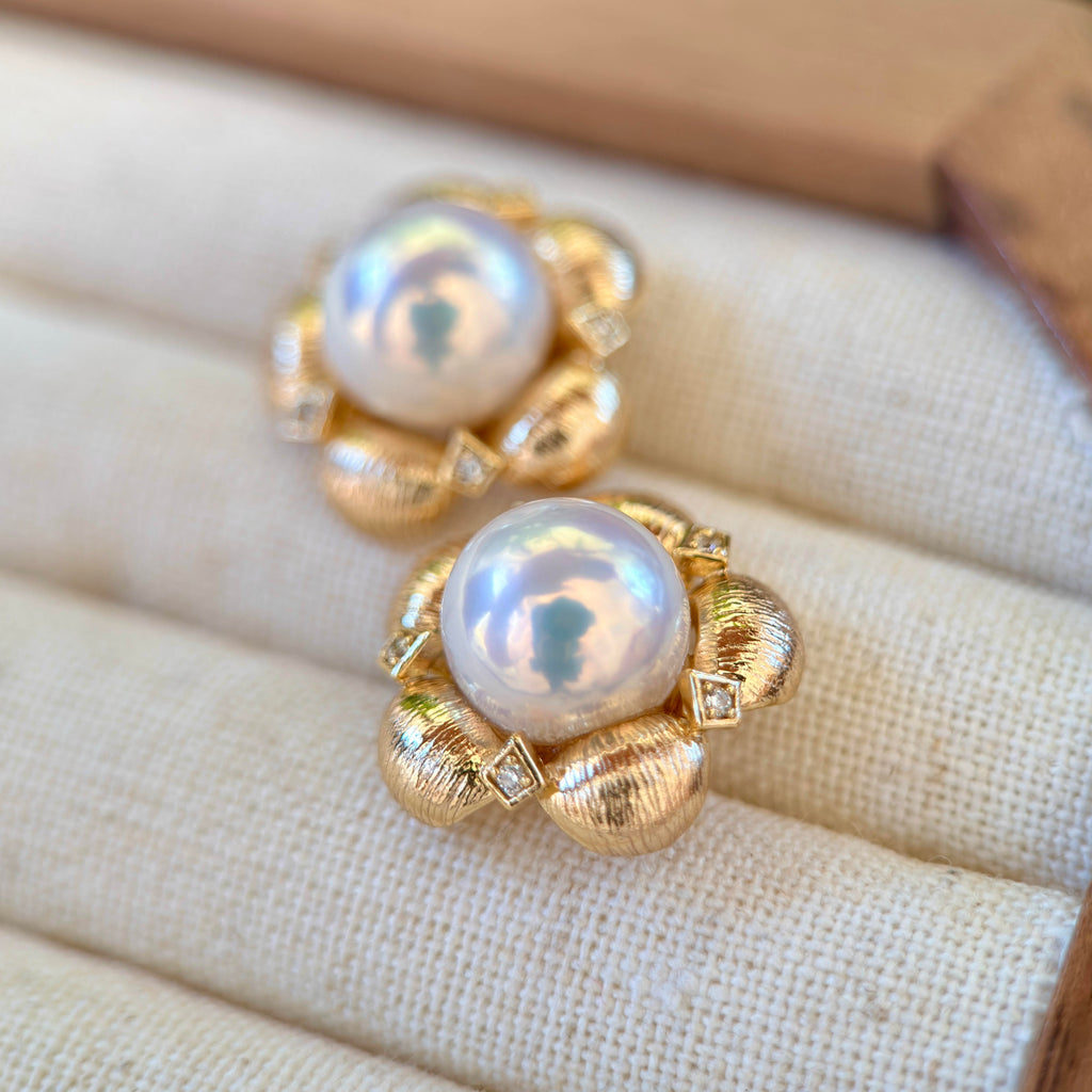 A pair of gold flower-shaped earrings with large iridescent white pearls and small clear stones, displayed on a cream fabric jewelry surface.