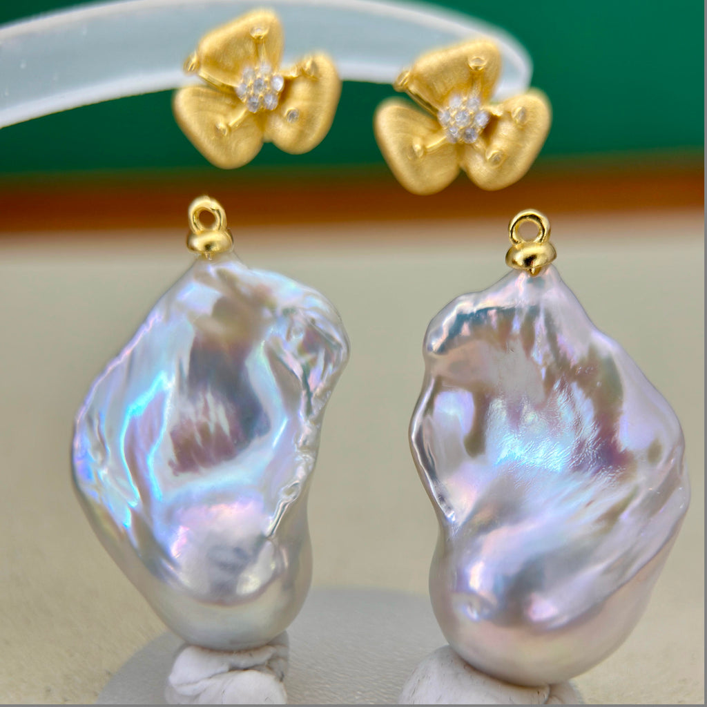 A pair of baroque pearl drop earrings with gold flower-shaped posts set with small clear stones, displayed on a neutral surface with a blurred green and brown background.