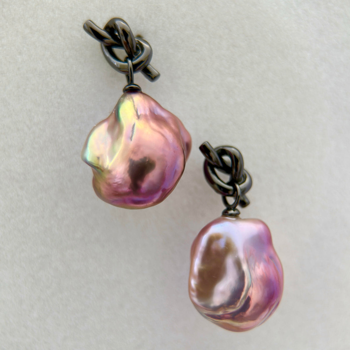 A pair of baroque pearl drop earrings with iridescent pink, purple, and green hues, suspended from dark oxidized metal jump rings on a light gray background.