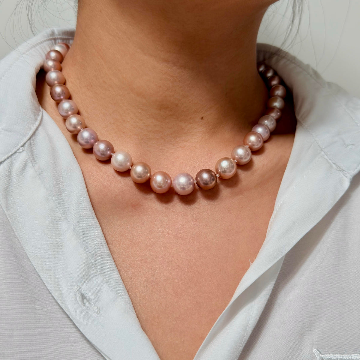 A close-up of a person wearing a single-strand pearl necklace with lustrous pinkish-white beads, paired with a white collared shirt.