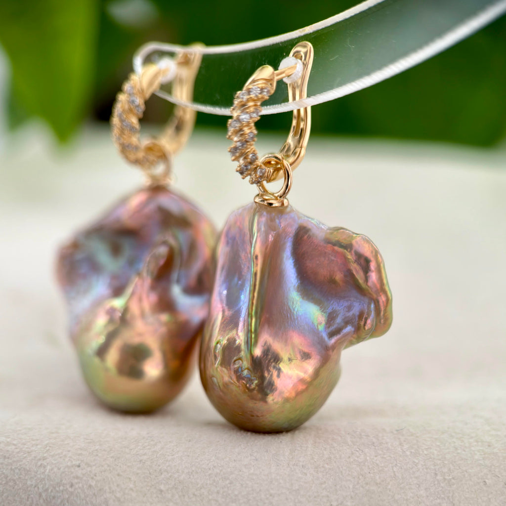 A pair of gold hoop earrings with large, baroque-shaped iridescent freshwater pearls, displayed on a soft beige surface with a blurred green background.