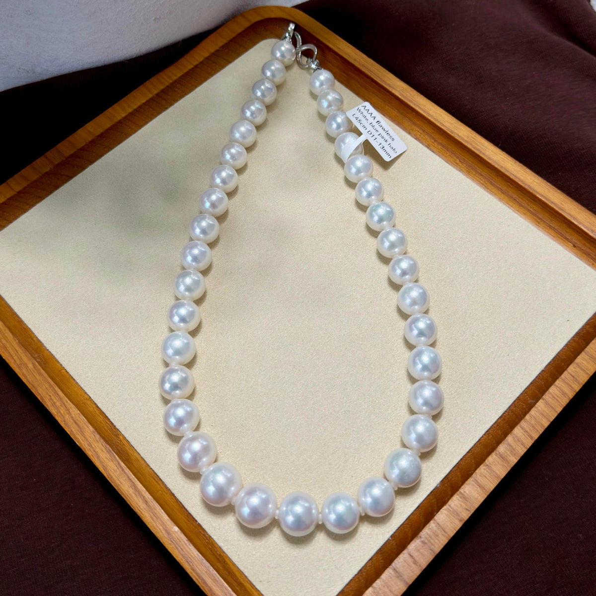 A strand of white freshwater pearls arranged in an oval shape on a beige display cushion inside a wooden box, with a small paper tag attached near the clasp.