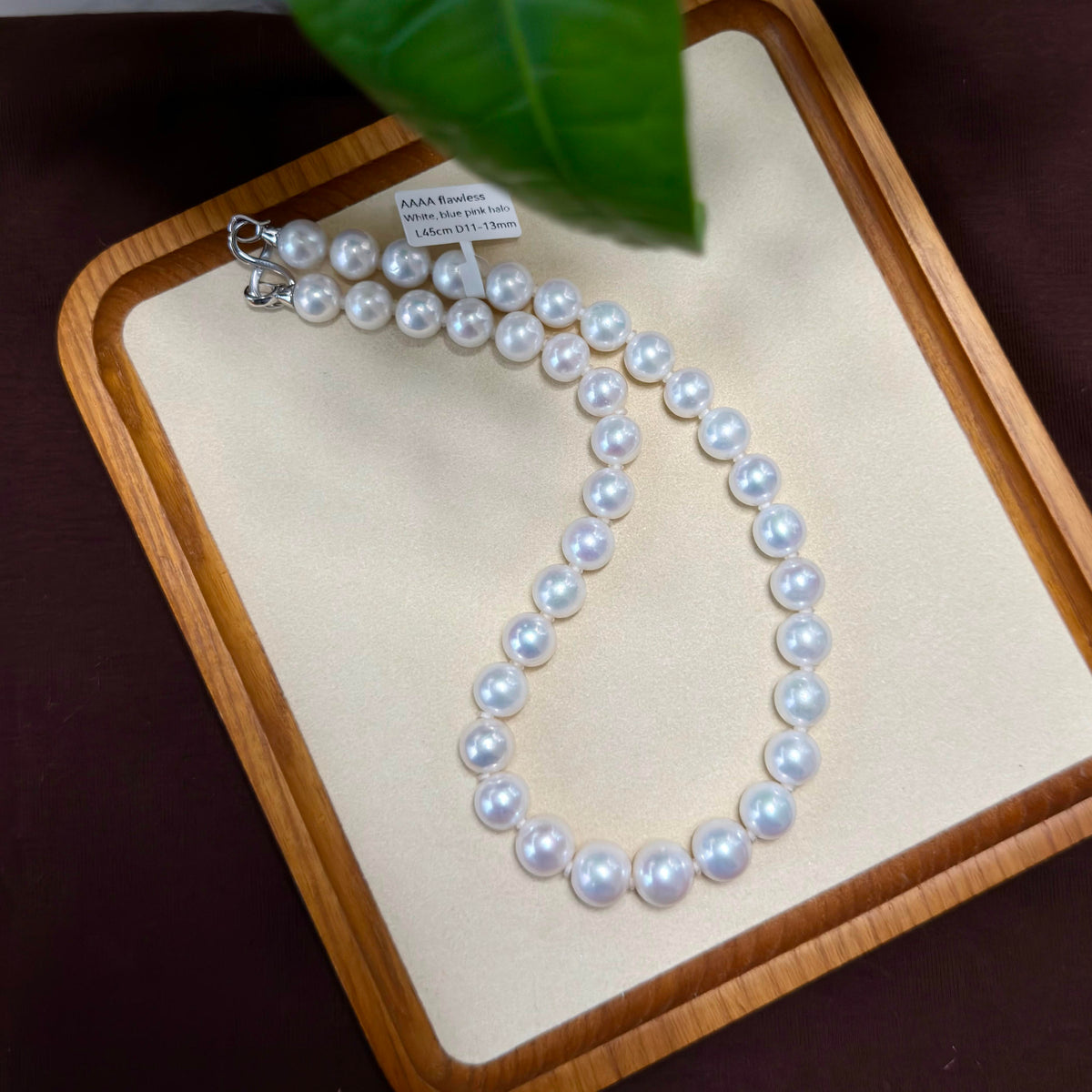 A strand of white freshwater pearl beads with a silver clasp, displayed on a cream-colored jewelry tray with a wooden border and a green leaf in the background.