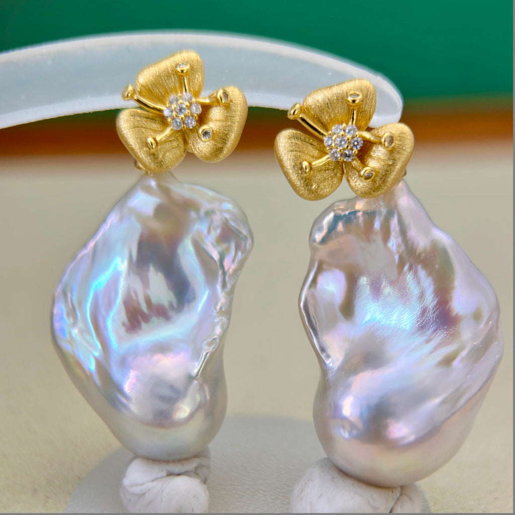 A pair of gold flower-shaped stud earrings with baroque pearl drops, displayed on a jewelry stand against a soft-focus background.