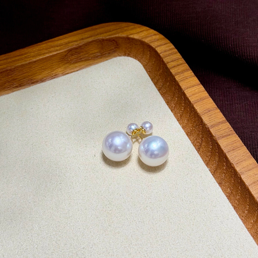 A pair of pearl stud earrings with small gold post backings, resting on a beige textured jewelry tray inside a wooden frame.