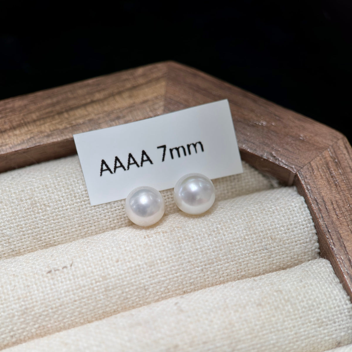 A pair of round white pearl stud earrings displayed on a beige fabric jewelry tray inside a wooden box, with a small card reading 'AAAA 7mm' placed above them.
