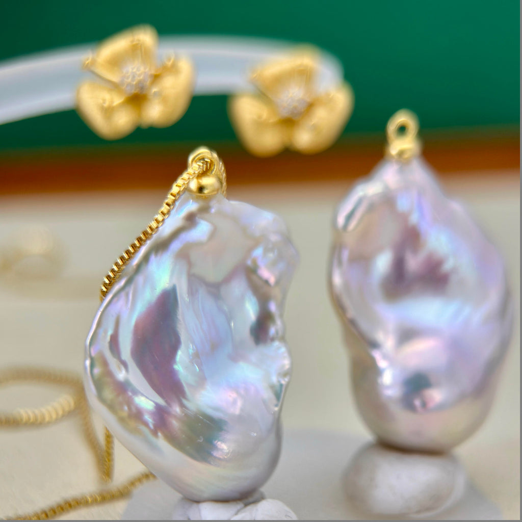 A close-up product shot showing a baroque pearl pendant necklace and matching baroque pearl stud earrings with gold settings, displayed on a light surface with a blurred green and brown background.