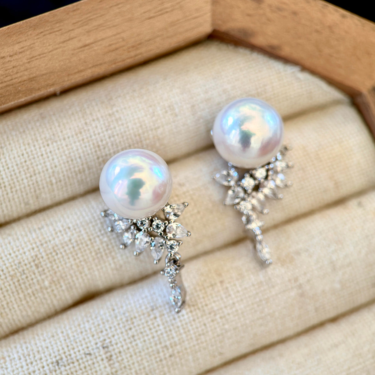 A pair of pearl and crystal drop earrings displayed on a beige fabric-lined jewelry box. Each earring features a round, lustrous white pearl set in a silver-toned metal cluster adorned with clear crystals.