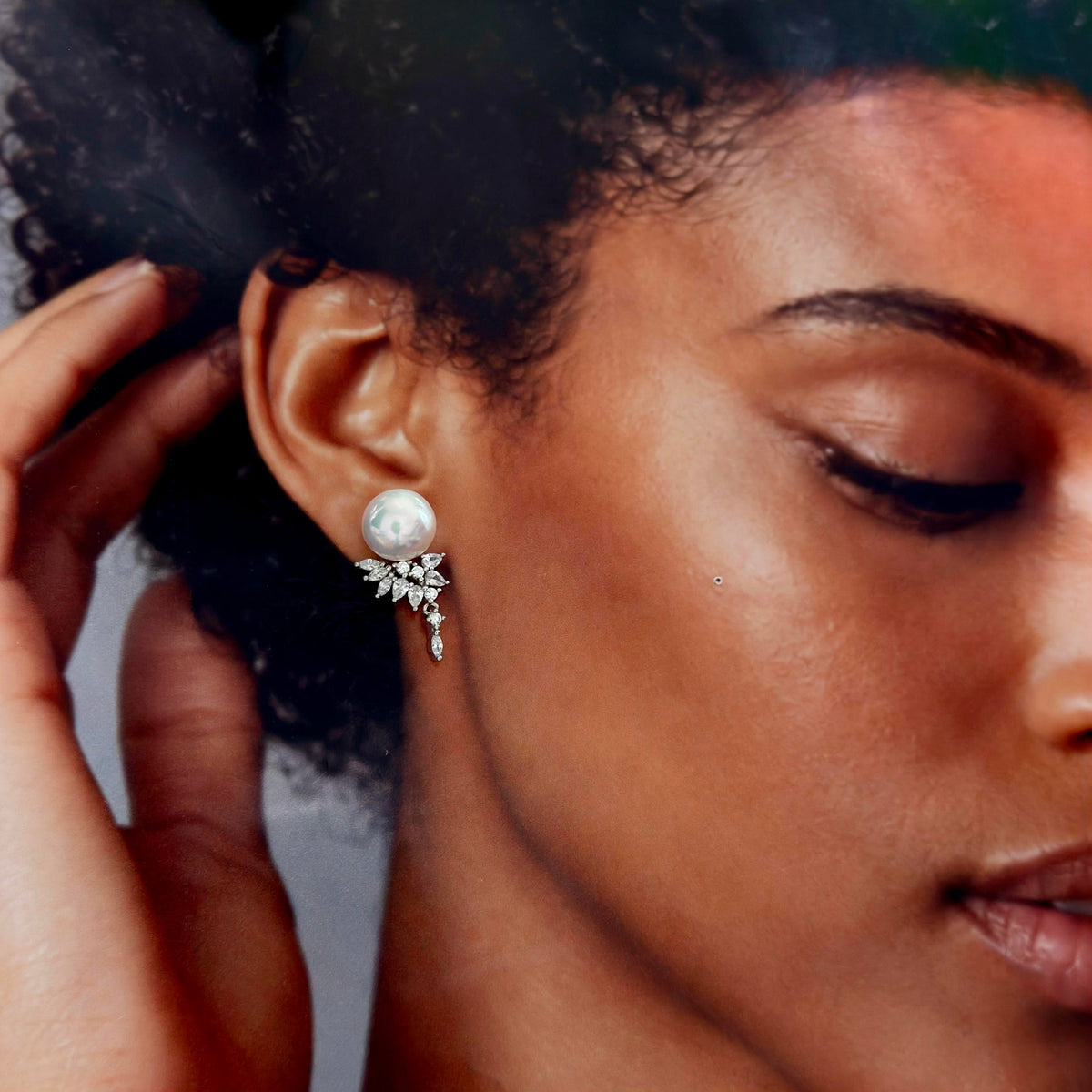 A close-up of a woman wearing a silver leaf-shaped drop earring with a white pearl and clear crystals, with her hand near her hair.