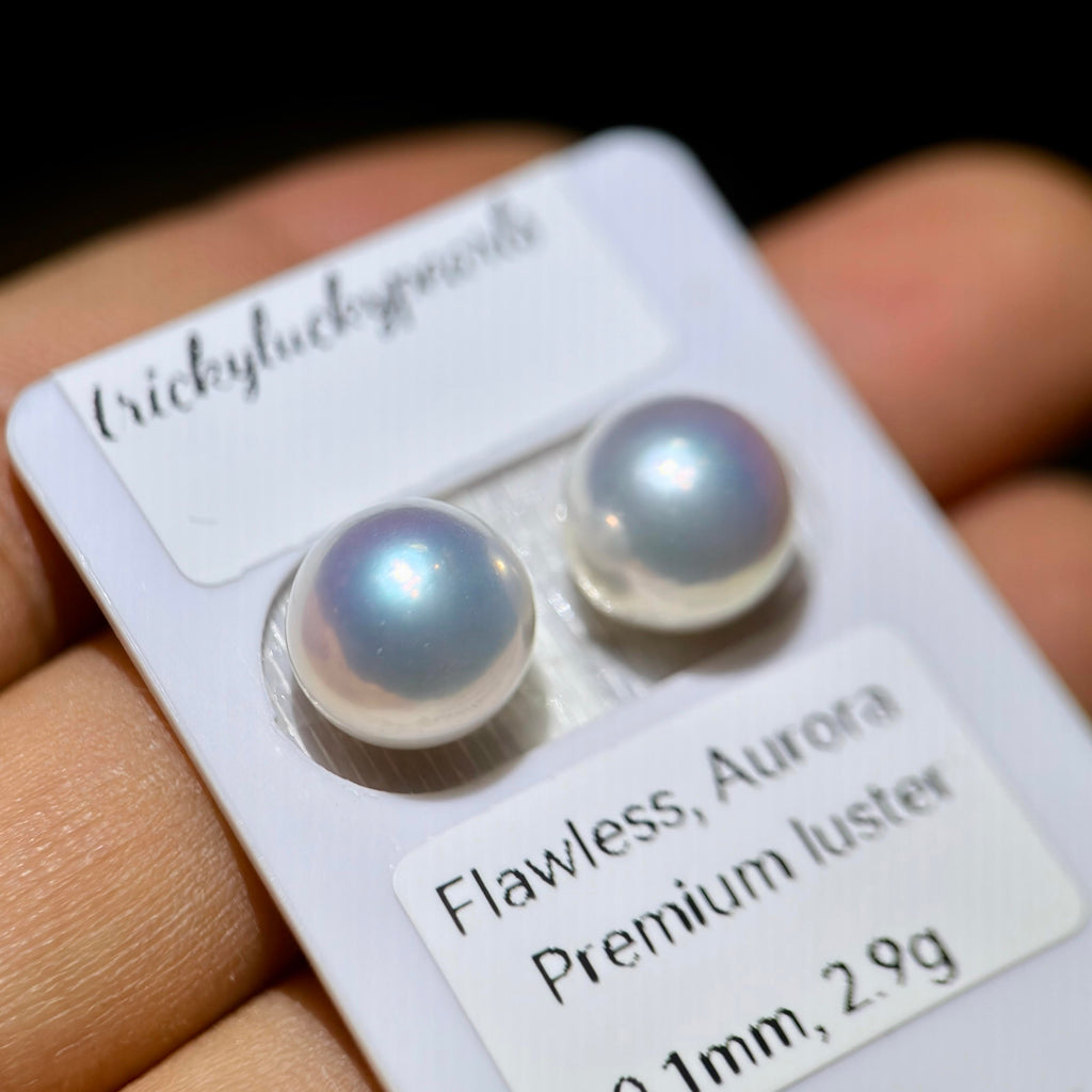 A close-up of a hand holding a white display card with two round pearl stud earrings. The card includes printed labels reading 'trickyluckgravel' and 'Flawless, Aurora Premium luster'.