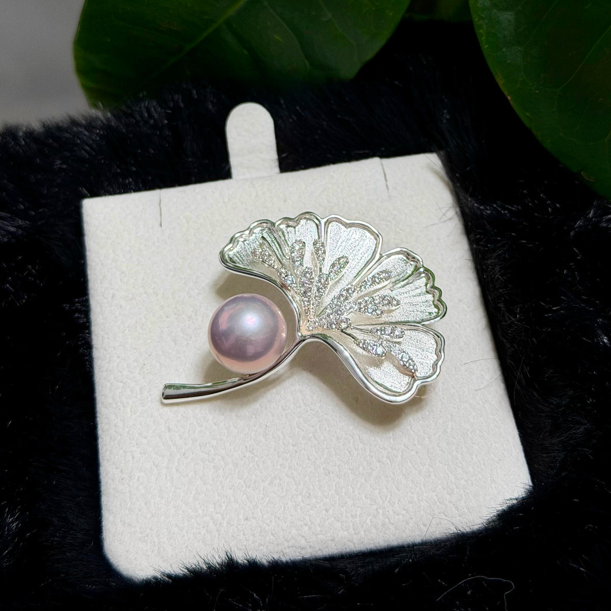 A silver ginkgo leaf brooch with a pink pearl accent, displayed on a white jewelry card over a black textured background with green leaves.