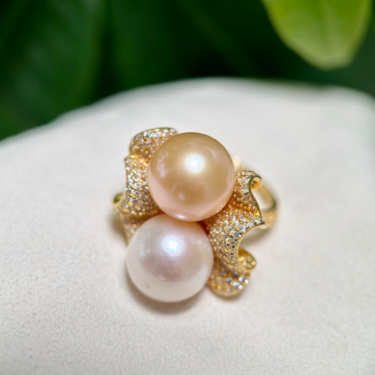 A gold ring featuring two large round pearl-like beads, one warm champagne beige and one soft white, set in a pavé diamond floral design, resting on a white textured surface with a blurred green background.