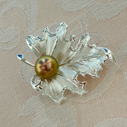 A silver-toned leaf-shaped brooch with a central round olive-green pearl, displayed on a textured beige fabric background.