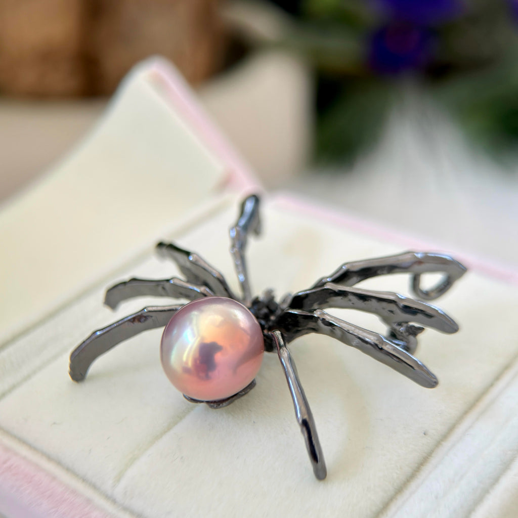 A spider-shaped ring with a pink iridescent pearl and metallic silver legs, displayed on a cream-colored jewelry box.