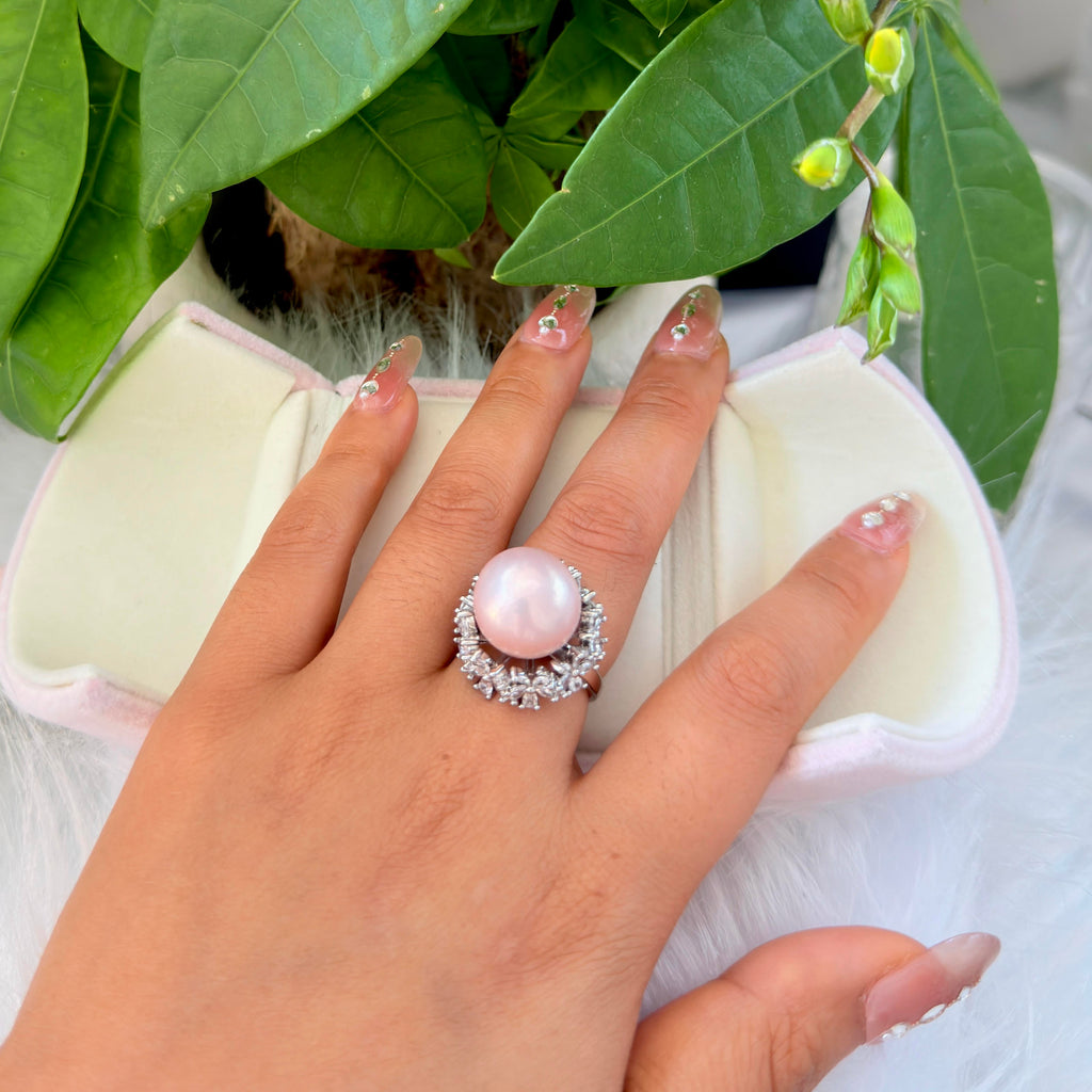 A close-up of a hand wearing a ring with a large round pale pink pearl-like center stone surrounded by small clear stones, resting over an open cream jewelry box with green leaves in the background.