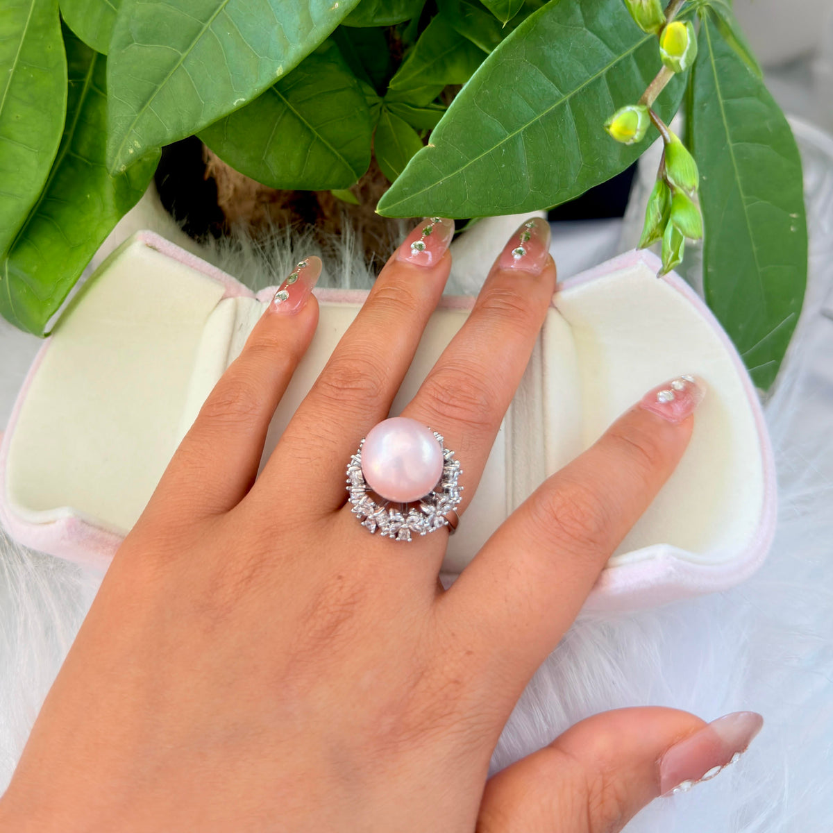 A close-up of a hand wearing a ring with a large round pale pink pearl-like center stone surrounded by small clear stones, resting over an open cream jewelry box with green leaves in the background.