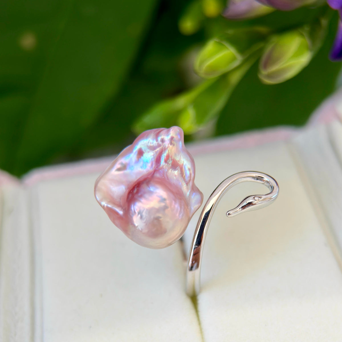 A baroque pearl ring with a silver hook setting, displayed in an open jewelry box with green leaves and purple flowers in the background.