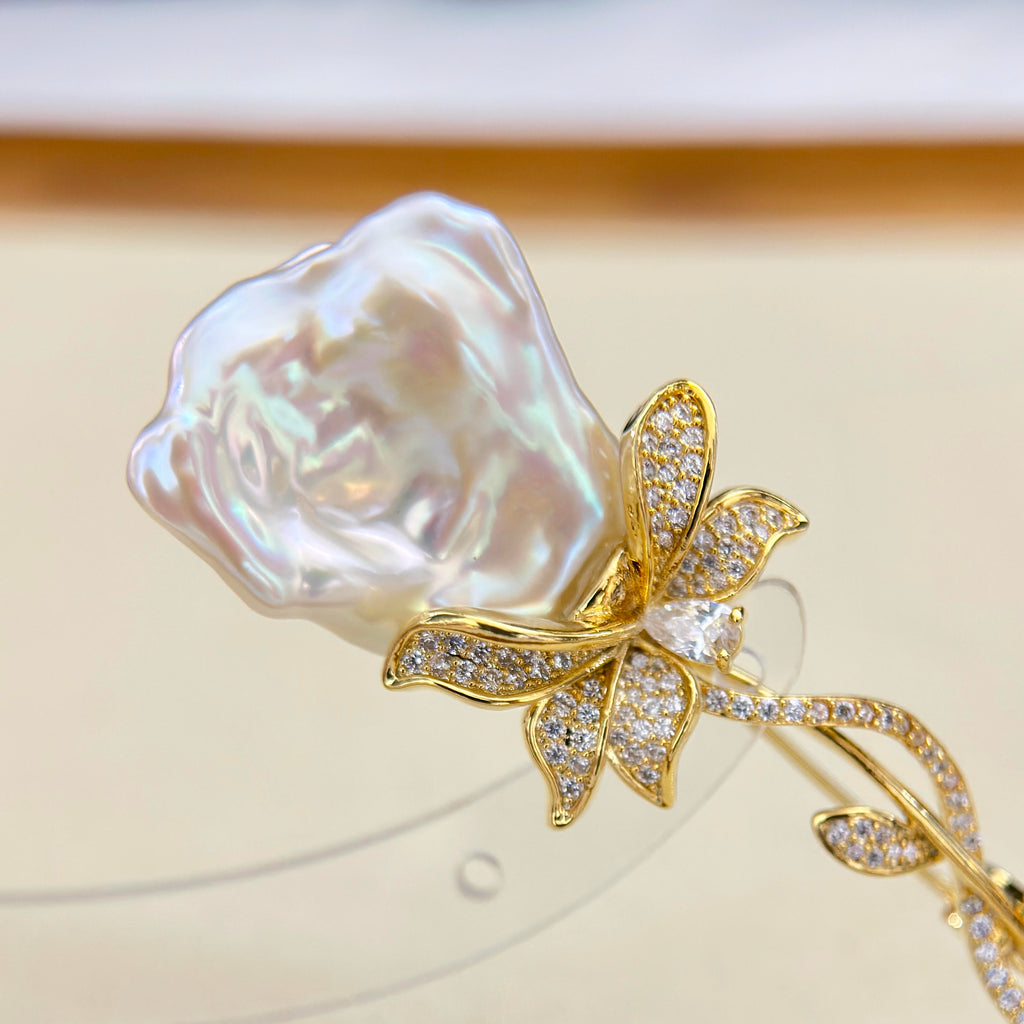 A close-up of a gold rose-shaped hair claw clip featuring a large, lustrous iridescent pearl flower head and a bow and stem decorated with small clear crystals, displayed on a clear stand against a soft beige background.