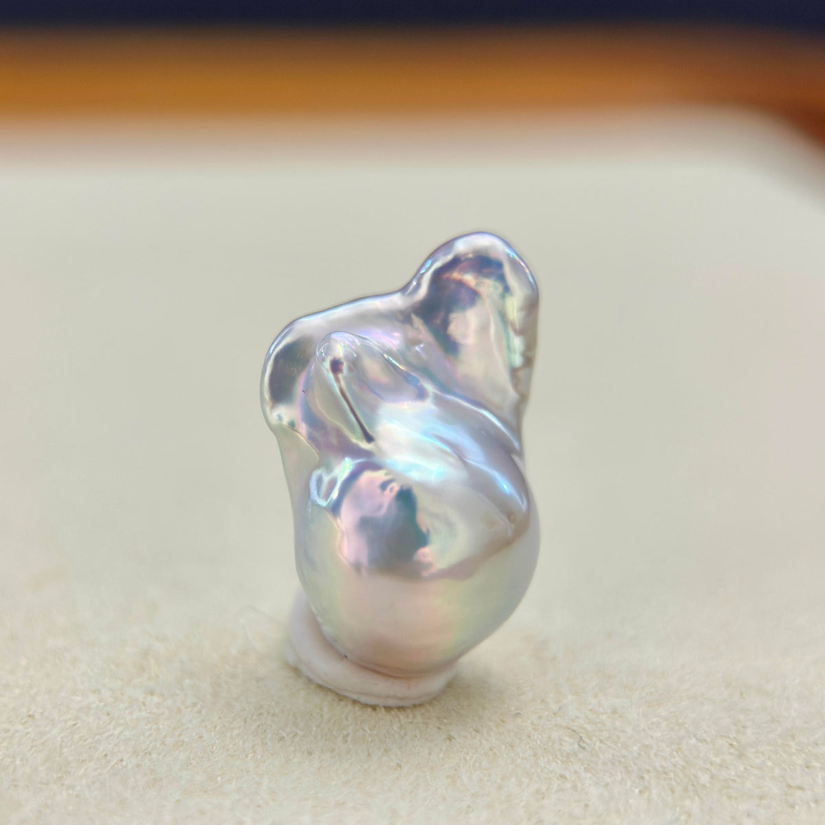 A single irregular baroque-style pearl with iridescent silver, blue, and pink highlights, photographed standing upright on a soft beige surface.