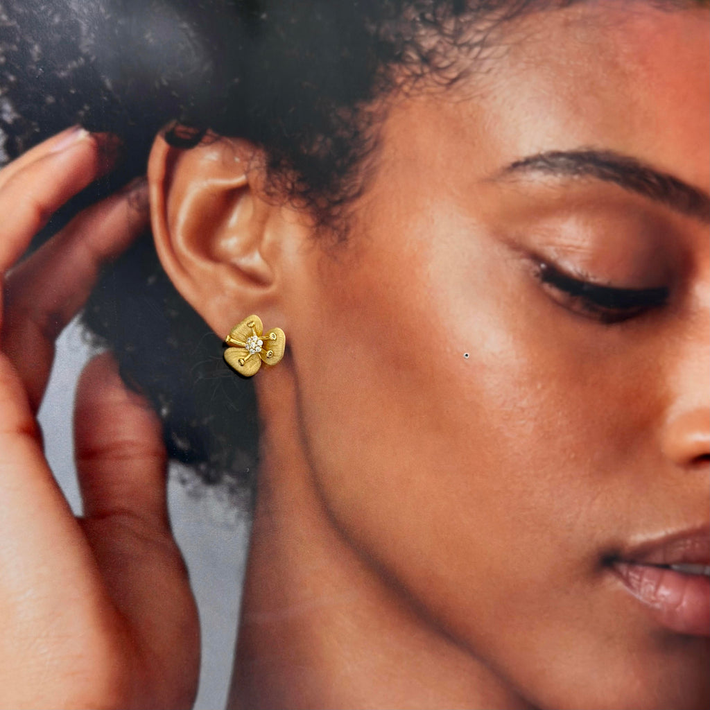 A close-up lifestyle shot of a woman wearing a small yellow gold flower-shaped stud earring with a clear central gemstone. Her hand is gently touching her hair, and the background is a soft, out-of-focus gray.