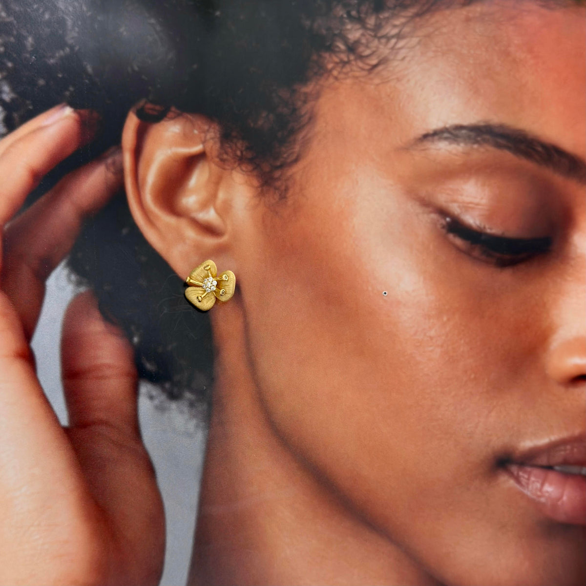 A close-up lifestyle shot of a woman wearing a small yellow gold flower-shaped stud earring with a clear central gemstone. Her hand is gently touching her hair, and the background is a soft, out-of-focus gray.