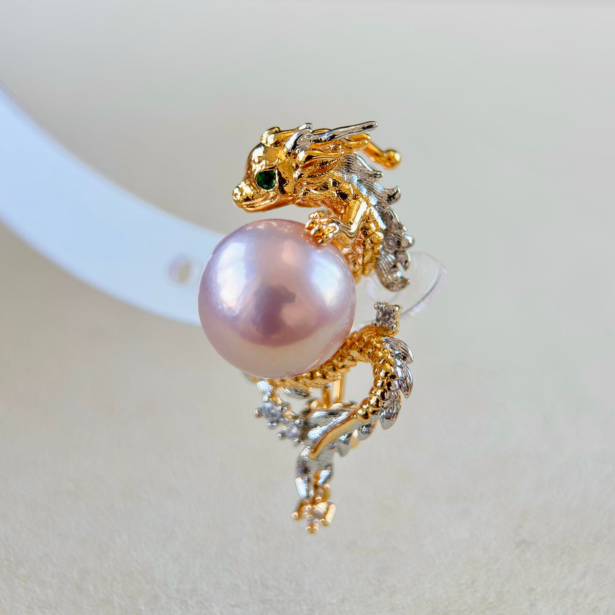 A gold dragon-shaped ring with a large pink pearl and small green and clear gemstone accents, displayed on a soft cream background.