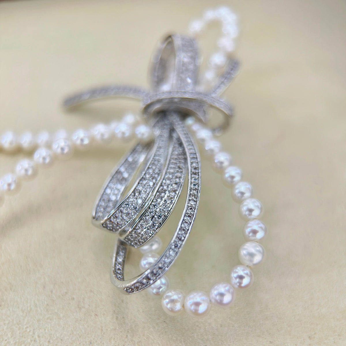 A silver-toned bow-shaped pendant embellished with pavé clear stones, draped over a strand of white pearl beads on a soft beige background.