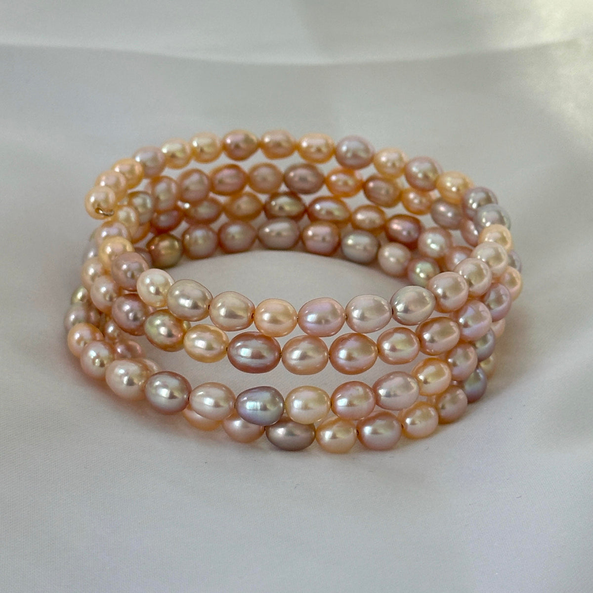 A multi-strand wrap bracelet made of irregular, lustrous freshwater pearls in soft peach, pink, cream, and champagne tones, arranged on a light fabric background.