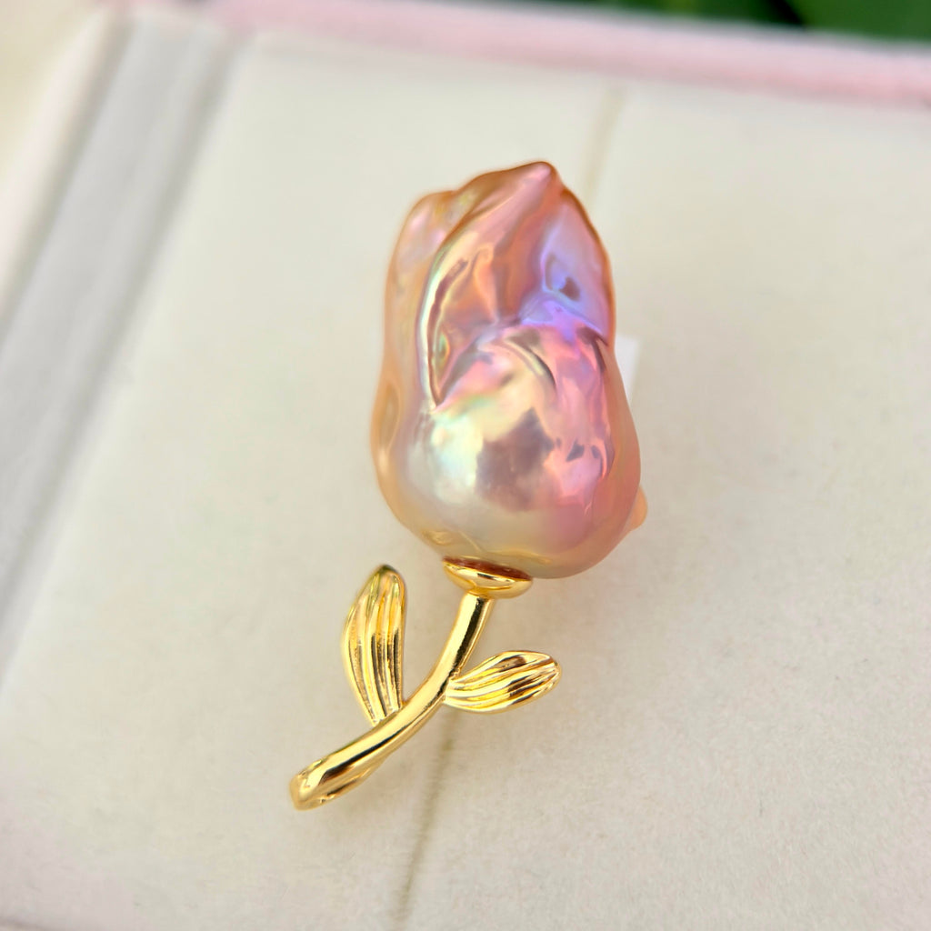 A rose-shaped baroque pearl brooch with a gold stem and leaves, resting on a light cream-colored jewelry box.