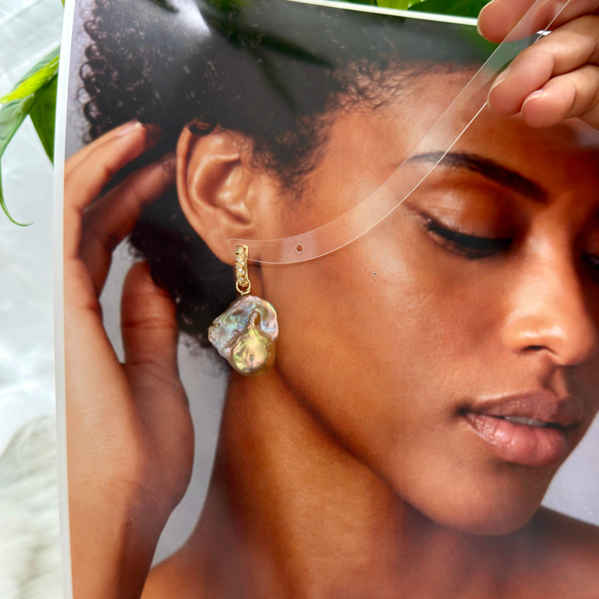 A close-up of a model wearing a gold earring with an irregular, iridescent shell-like drop pendant. The photo is taken from a side angle, showing the model's ear, hair, and hand adjusting the earring, with a clear plastic sheet partially covering the top of the frame.