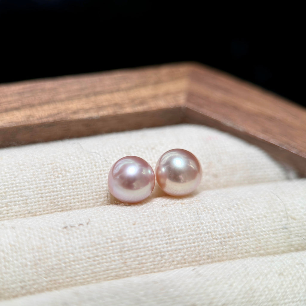 A pair of round, lustrous pink freshwater pearls resting on a cream-colored textured fabric surface inside a wooden jewelry box.