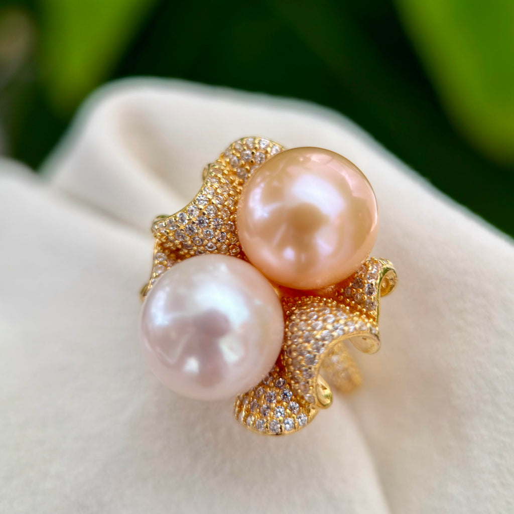 A gold ring featuring two large, lustrous pinkish pearls and pavé-set clear stones, displayed on a soft white fabric surface with a blurred green background.