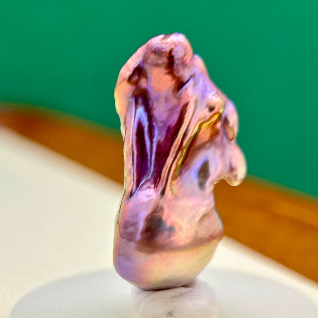 A polished, organic-shaped iridescent stone or pearl-like cabochon with swirling purple, pink, peach, and gold tones, displayed upright on a small white stand against a green and brown background.