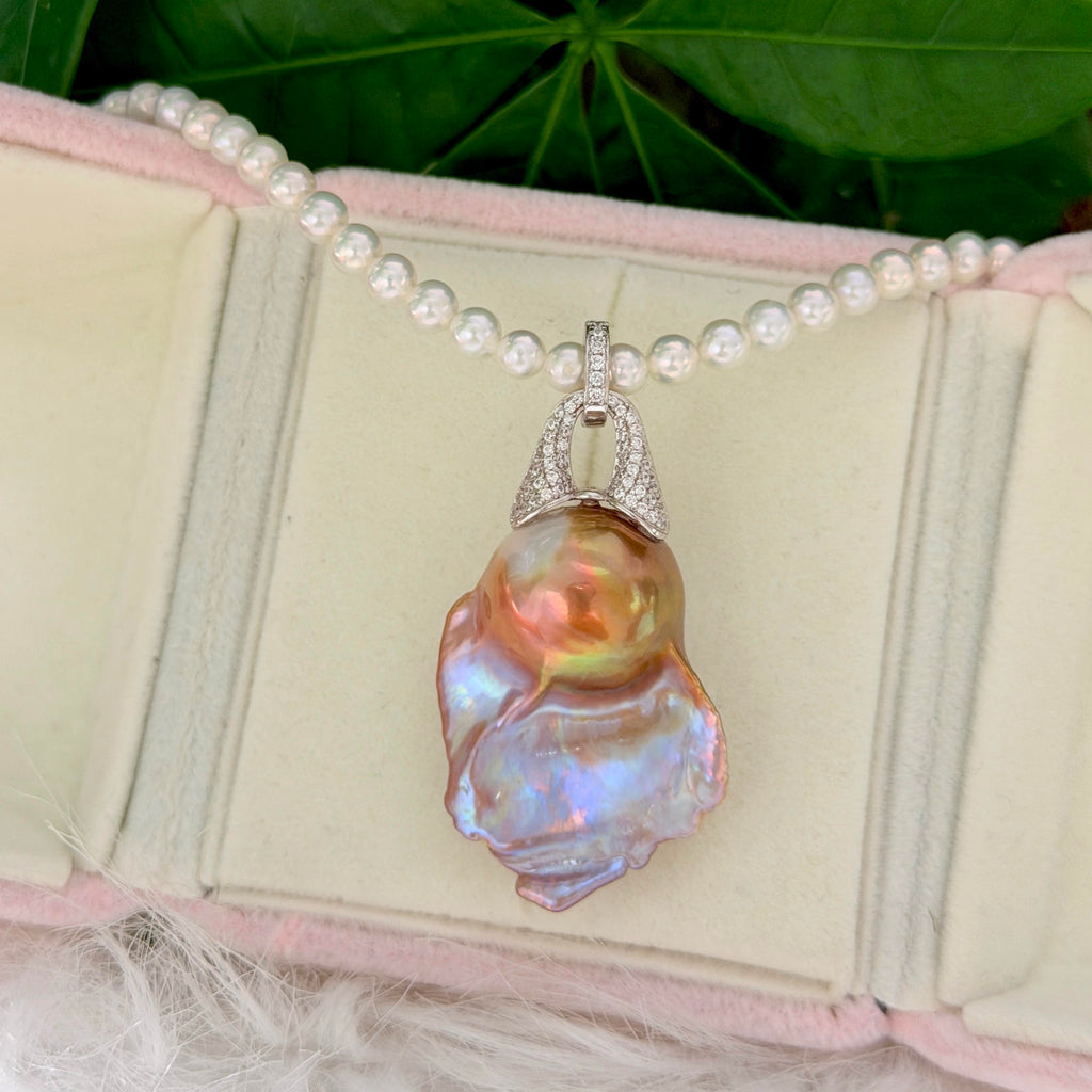 A pearl necklace with a large, iridescent baroque pearl pendant, displayed on a pink-lined jewelry box with green leaves in the background.