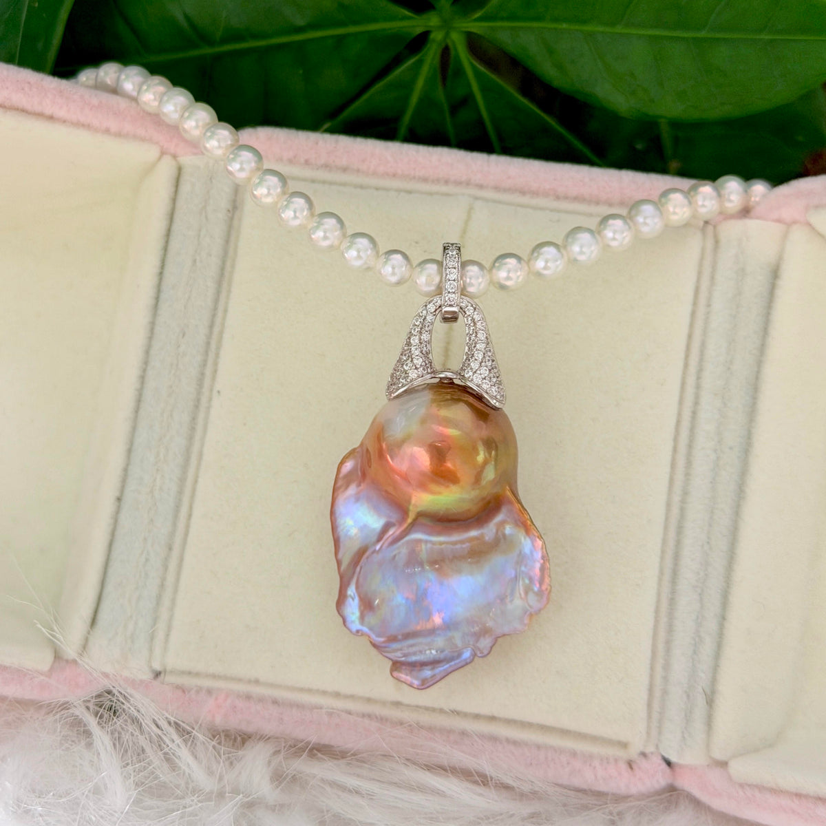 A pearl necklace with a large, iridescent baroque pearl pendant, displayed on a pink-lined jewelry box with green leaves in the background.
