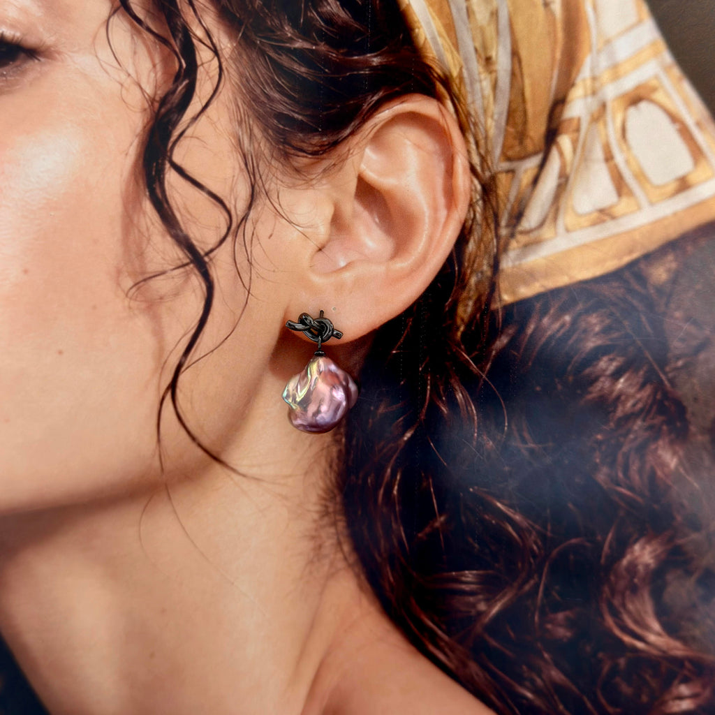 A close-up of a woman's ear wearing a dangling earring with a black enamel cross top and a teardrop-shaped iridescent purple charm.
