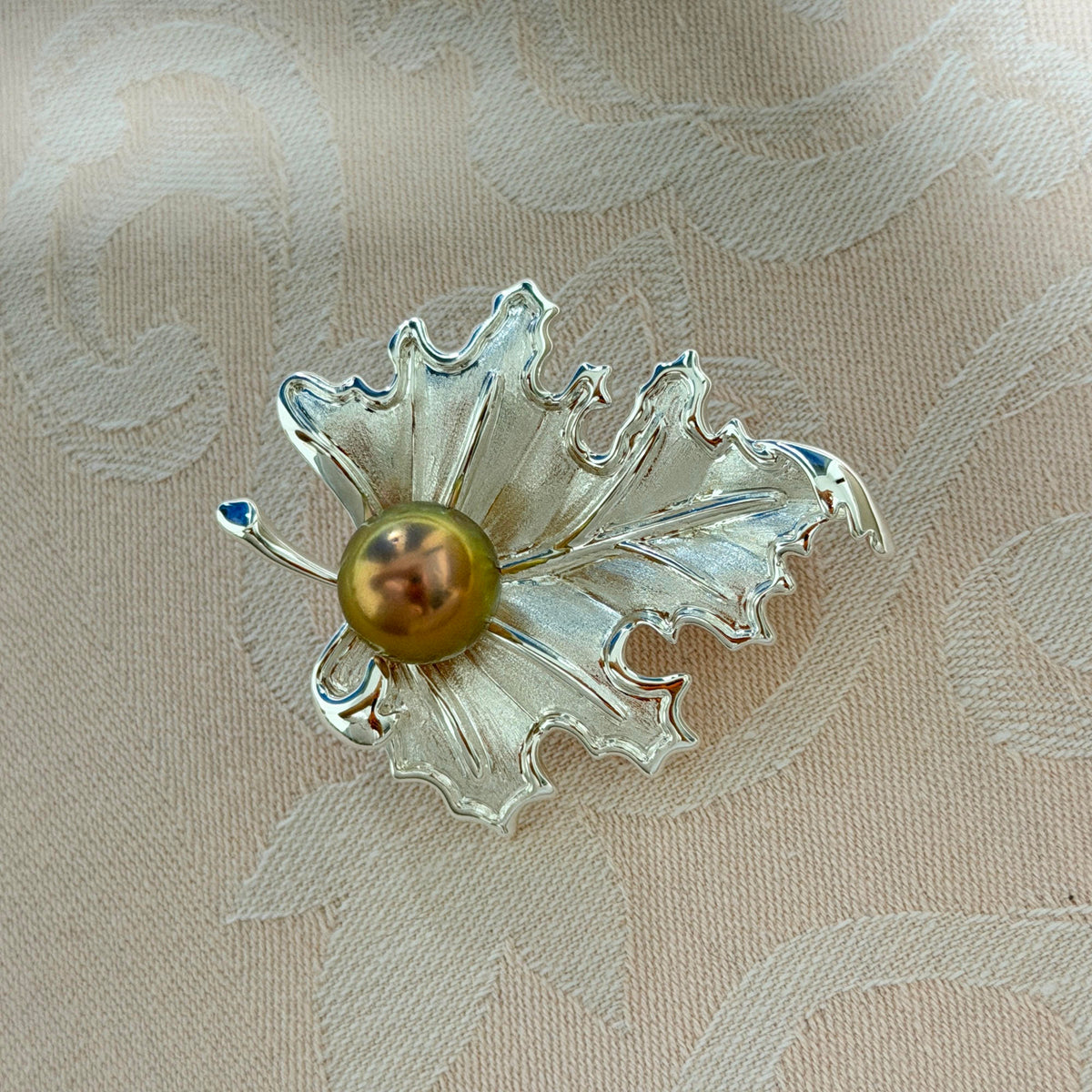 A silver-toned leaf-shaped brooch with a central round olive-green pearl, resting on a beige patterned fabric background.