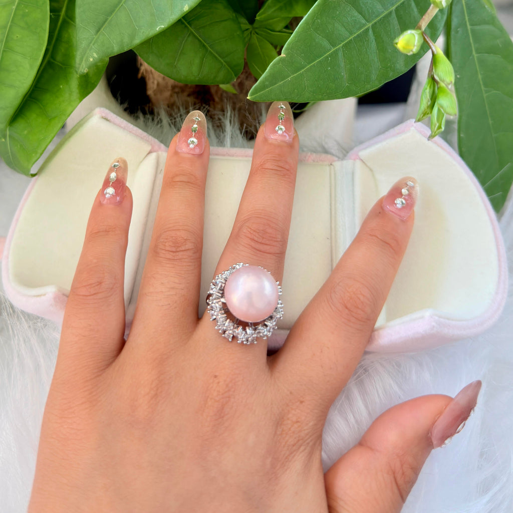 A hand with decorated nails wears a silver ring featuring a large pale pink pearl surrounded by small clear stones. An open pink jewelry box and green leaves are visible in the background.