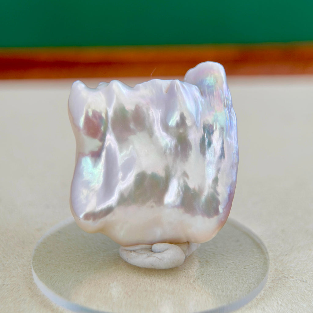 A large, irregular baroque pearl with a pearlescent white and iridescent lavender sheen, displayed on a small clear acrylic stand.