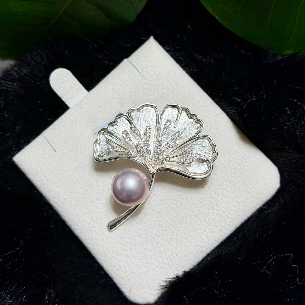 A silver ginkgo leaf brooch with a single pink pearl, displayed on a white square card over a black textured background with green leaves at the top.