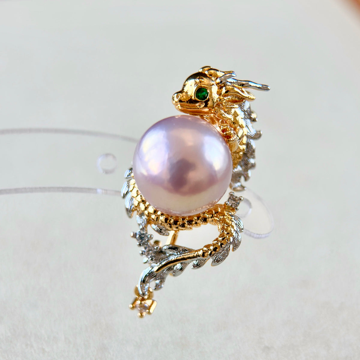 A dragon-shaped brooch featuring a large lustrous pearl, gold-toned metal, silver-toned accents, and a small green stone in the eye, photographed on a light neutral background.